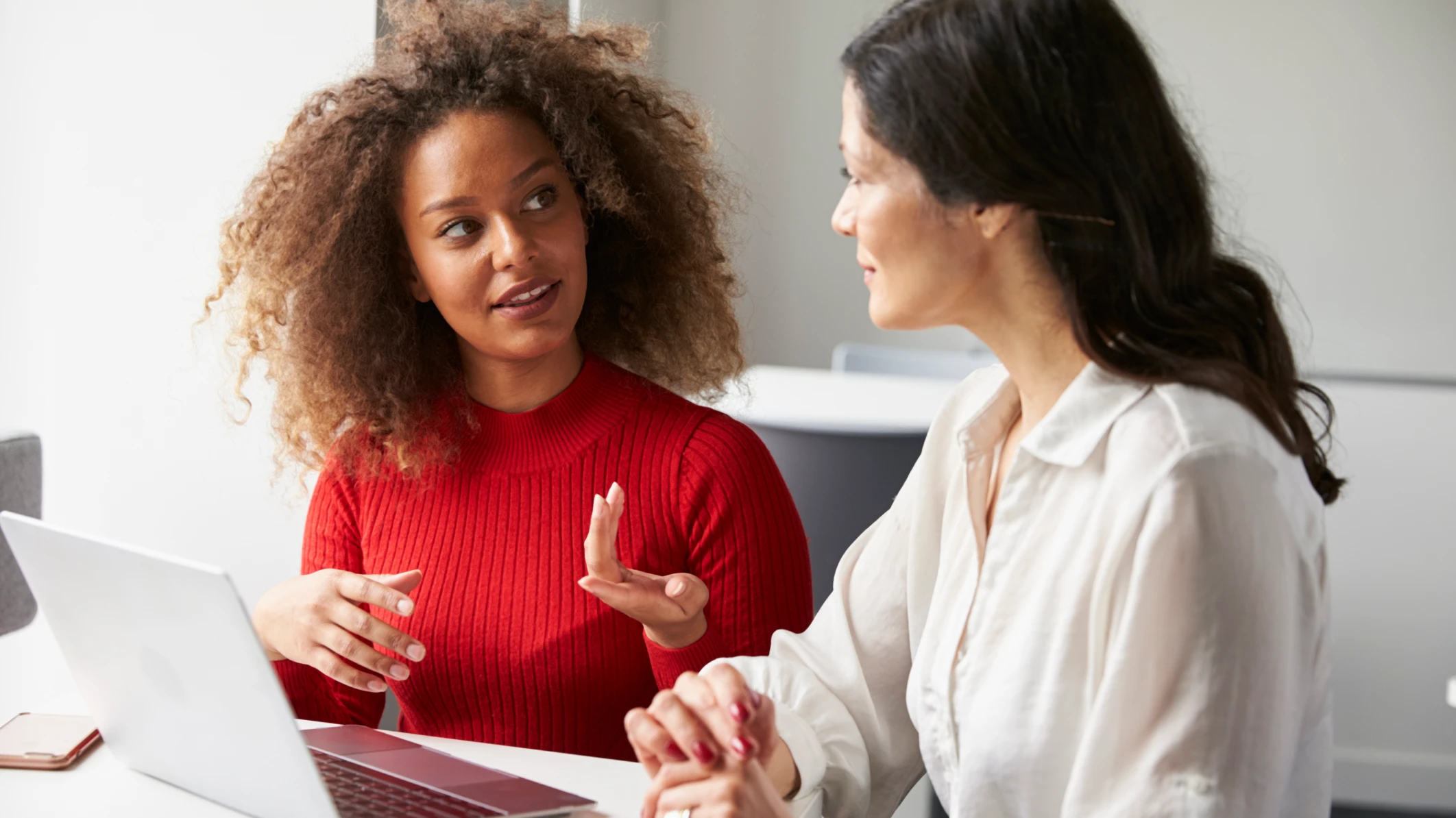 Black woman in red sweater is explaining something to a new employee: white woman in white blouse