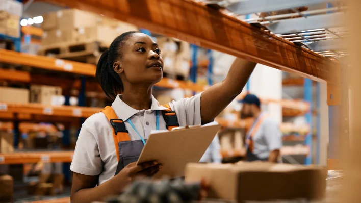 Mujer joven afroamericana con una carpeta en la mano alcanzando algo de un estante de almacén. Ella lleva un delantal de trabajo y una camisa blanca. 