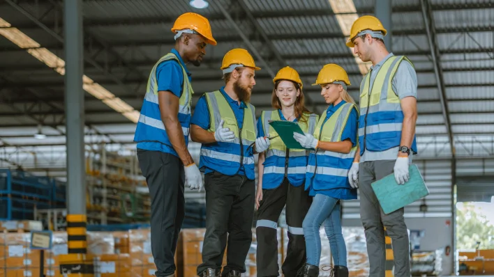 Una fila de 6 trabajadores multiculturales de una fábrica con cascos y guantes de trabajo. Todos miran a la cámara con el pulgar hacia arriba.