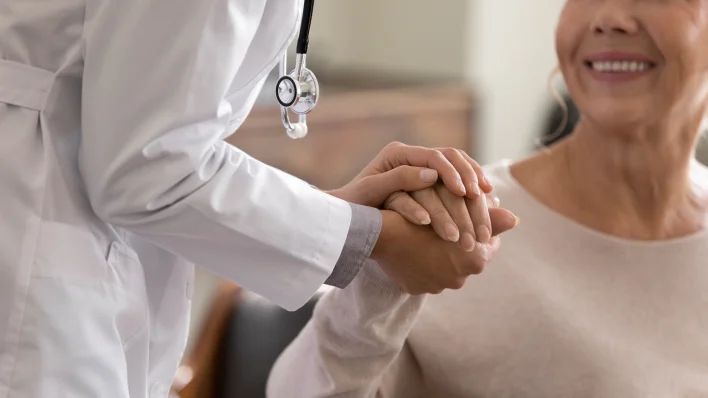 Entry level healthcare jobs: neck-down view of white-coated woman with stethoscope holding hands with a gray-haired woman. 