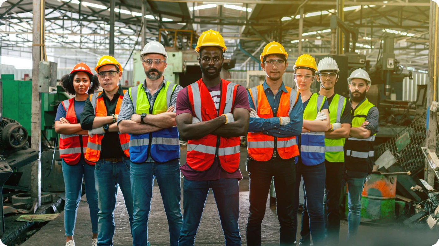 8 workers in safety vests, mixed ethnicities and sexes, stand facing the camera in a factory setting.